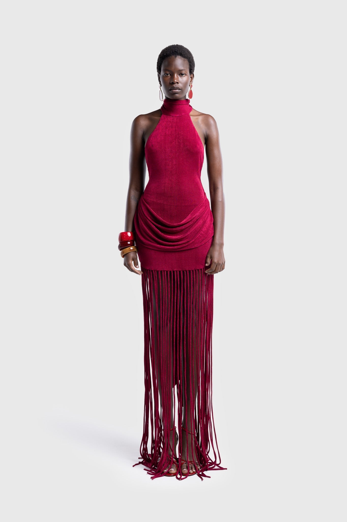 Full-length studio shot of a Black model wearing a sleeveless, high-neck halter dress in a deep red. The dress is form-fitting with ruching at the hips and features a dramatic, floor-length hem made of long, thick corded fringe.