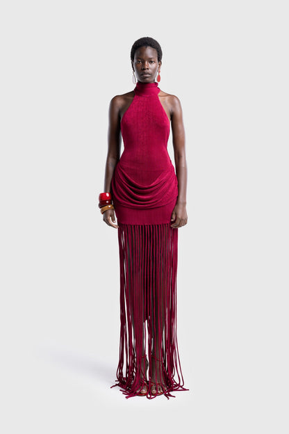 Full-length studio shot of a Black model wearing a sleeveless, high-neck halter dress in a deep red. The dress is form-fitting with ruching at the hips and features a dramatic, floor-length hem made of long, thick corded fringe.