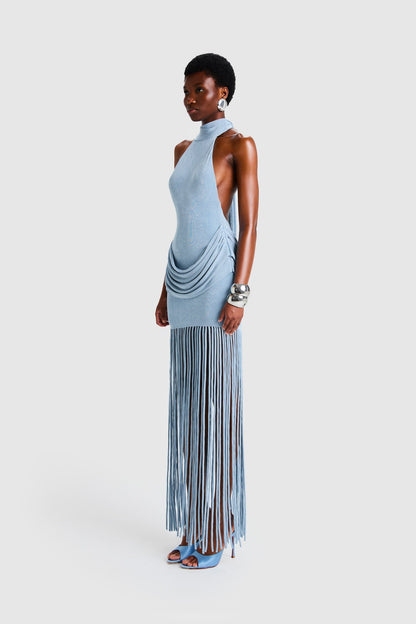 Full-length studio shot of a Black model wearing a sleeveless, high-neck halter dress in a deep red. The dress is form-fitting with ruching at the hips and features a dramatic, floor-length hem made of long, thick corded fringe.
