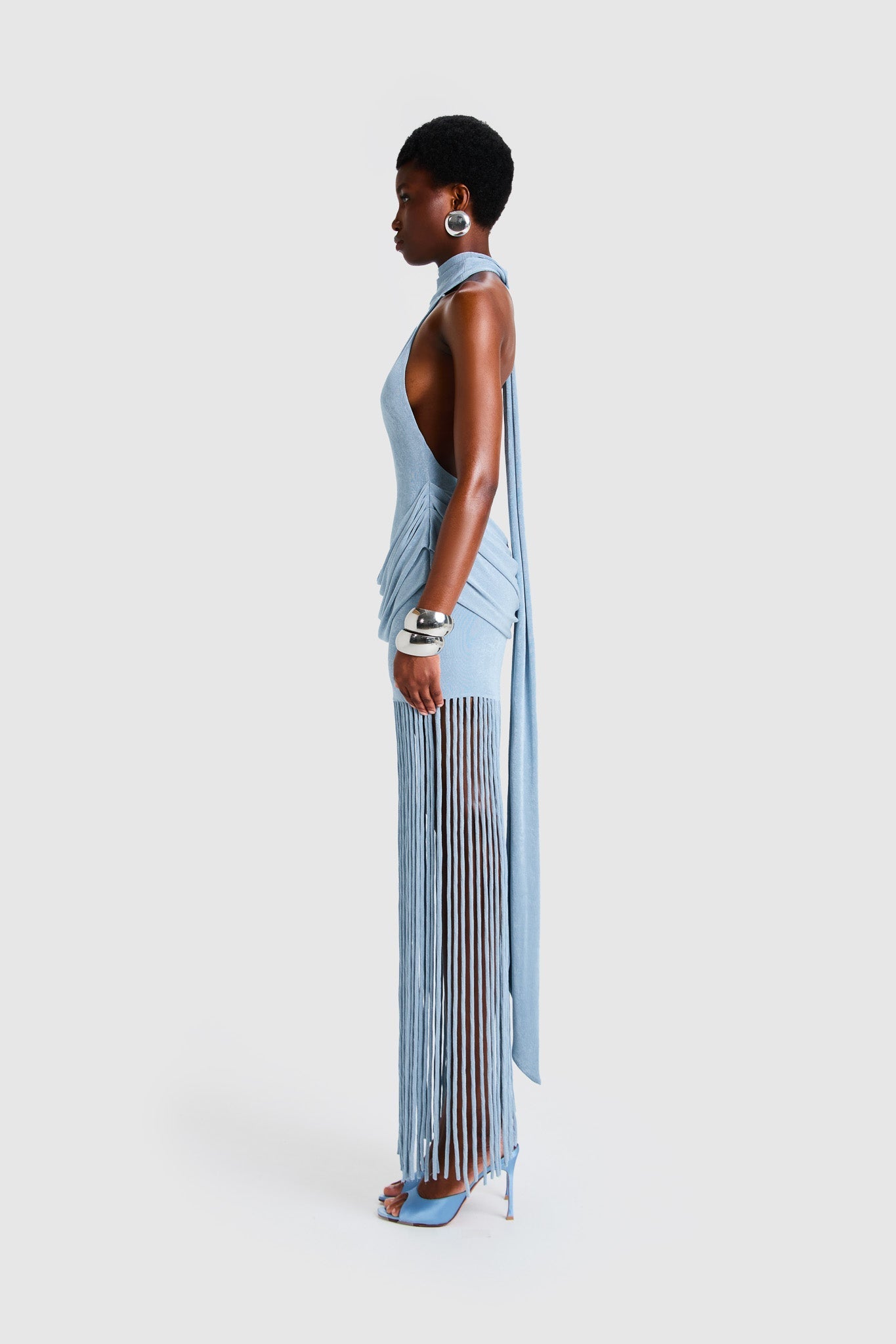 Full-length studio shot of a Black model wearing a sleeveless, high-neck halter dress in a deep red. The dress is form-fitting with ruching at the hips and features a dramatic, floor-length hem made of long, thick corded fringe.