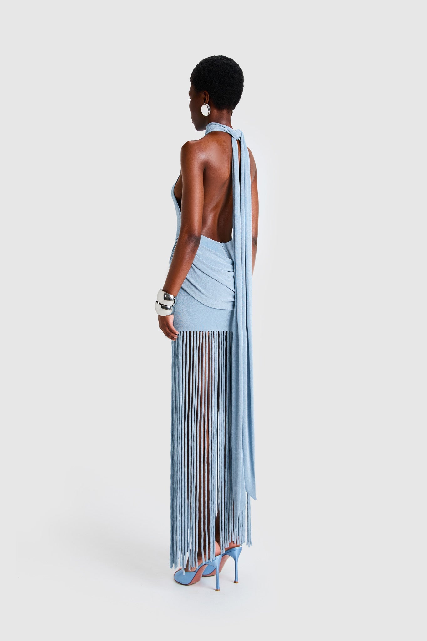 Full-length studio shot of a Black model wearing a sleeveless, high-neck halter dress in a deep red. The dress is form-fitting with ruching at the hips and features a dramatic, floor-length hem made of long, thick corded fringe.