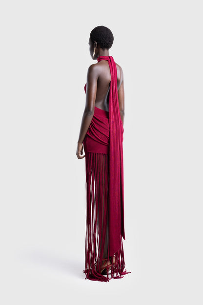 Full-length studio shot of a Black model wearing a sleeveless, high-neck halter dress in a deep red. The dress is form-fitting with ruching at the hips and features a dramatic, floor-length hem made of long, thick corded fringe.