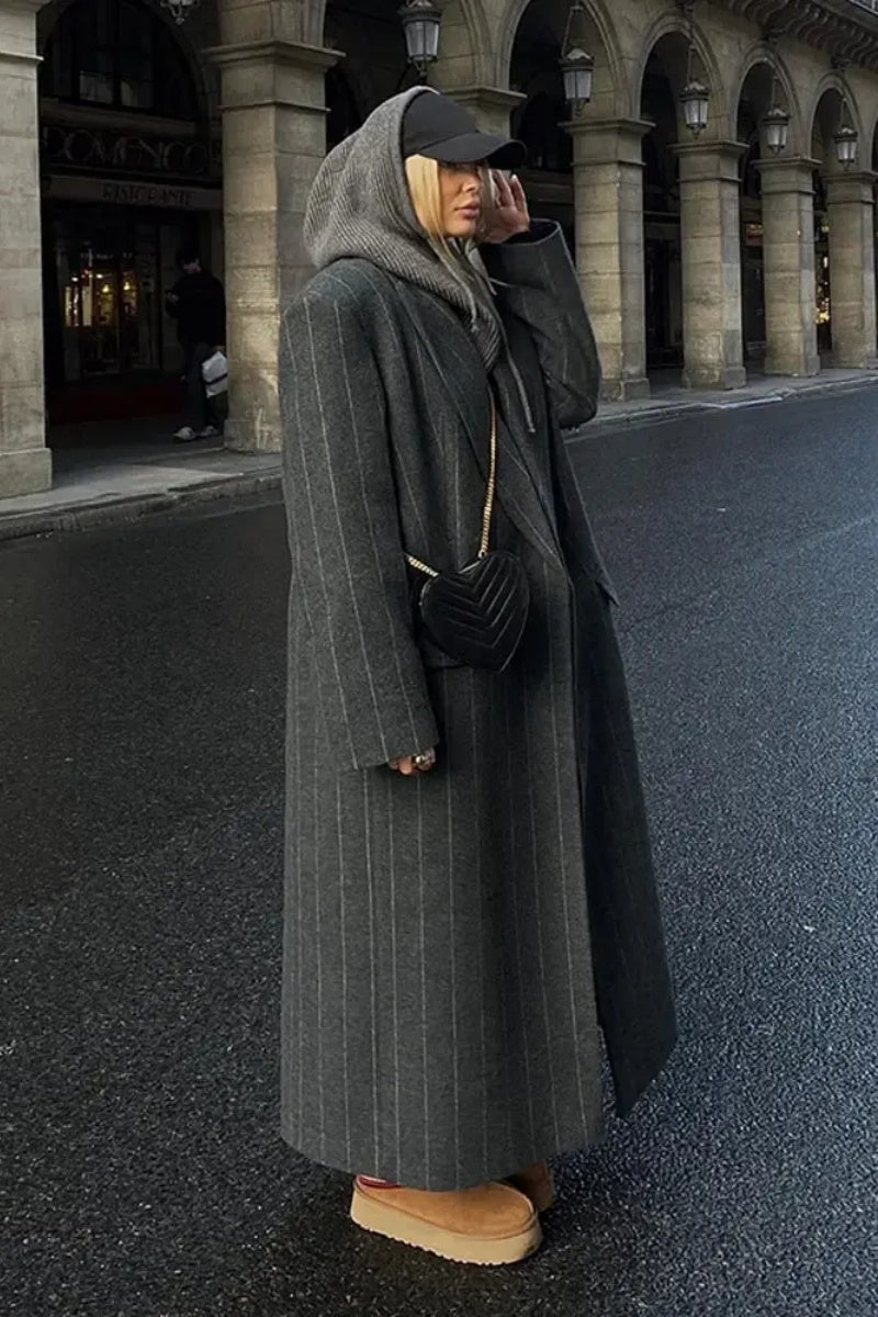 Grey - A woman walking on a city street wearing a dark grey, oversized maxi-length coat with subtle pinstripes, styled over dark jeans and a grey knit hood.