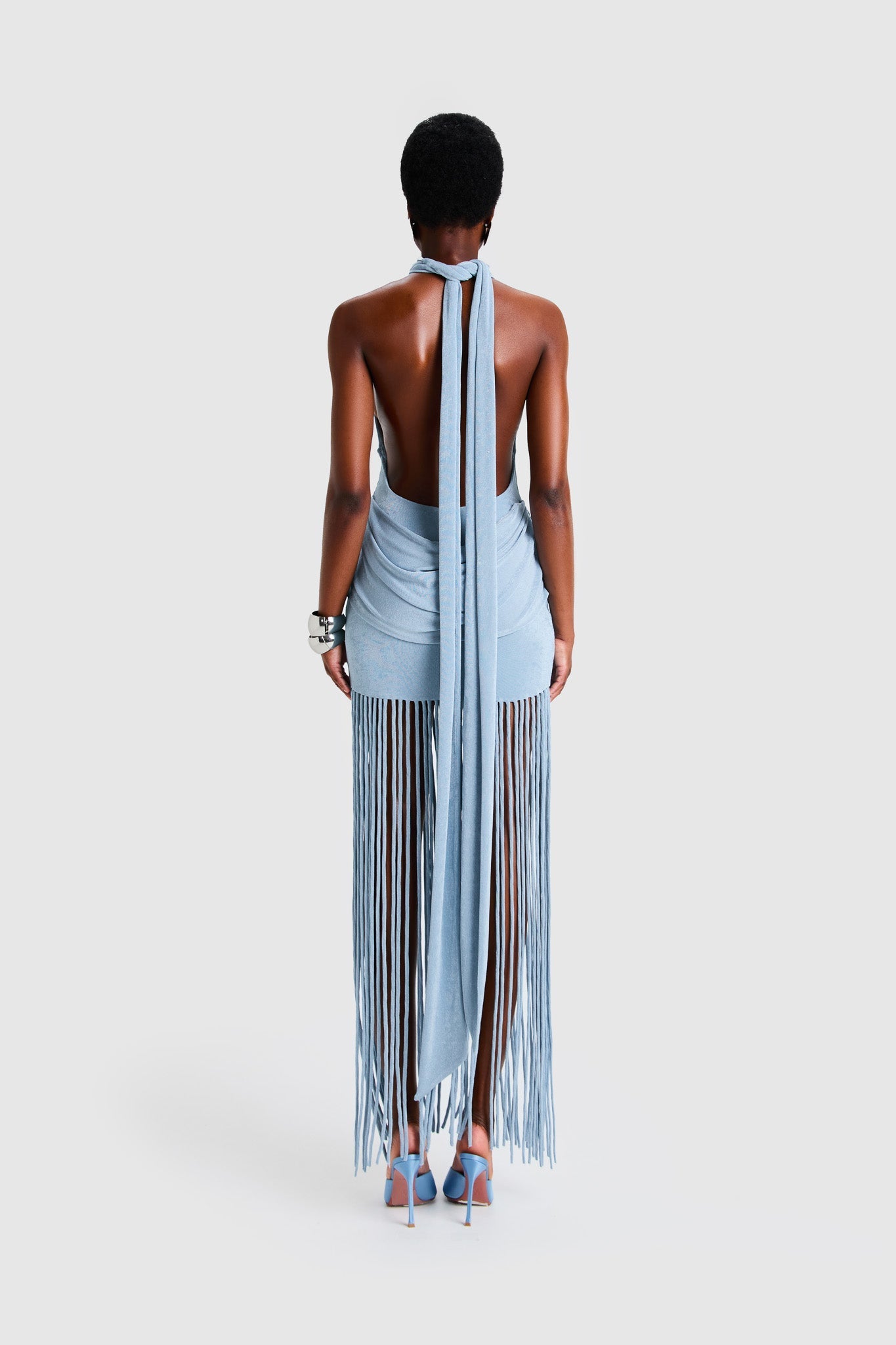 Full-length studio shot of a Black model wearing a sleeveless, high-neck halter dress in a deep red. The dress is form-fitting with ruching at the hips and features a dramatic, floor-length hem made of long, thick corded fringe.