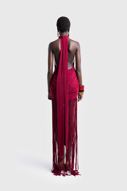 Full-length studio shot of a Black model wearing a sleeveless, high-neck halter dress in a deep red. The dress is form-fitting with ruching at the hips and features a dramatic, floor-length hem made of long, thick corded fringe.
