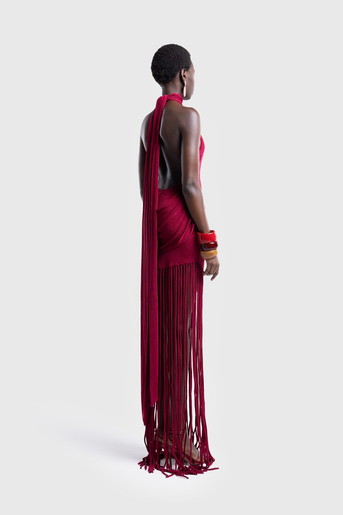 Full-length studio shot of a Black model wearing a sleeveless, high-neck halter dress in a deep red. The dress is form-fitting with ruching at the hips and features a dramatic, floor-length hem made of long, thick corded fringe.