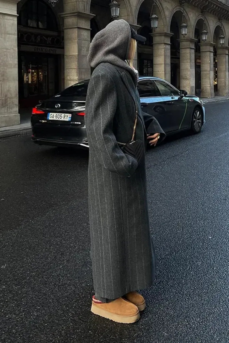 Grey - A woman walking on a city street wearing a dark grey, oversized maxi-length coat with subtle pinstripes, styled over dark jeans and a grey knit hood.
