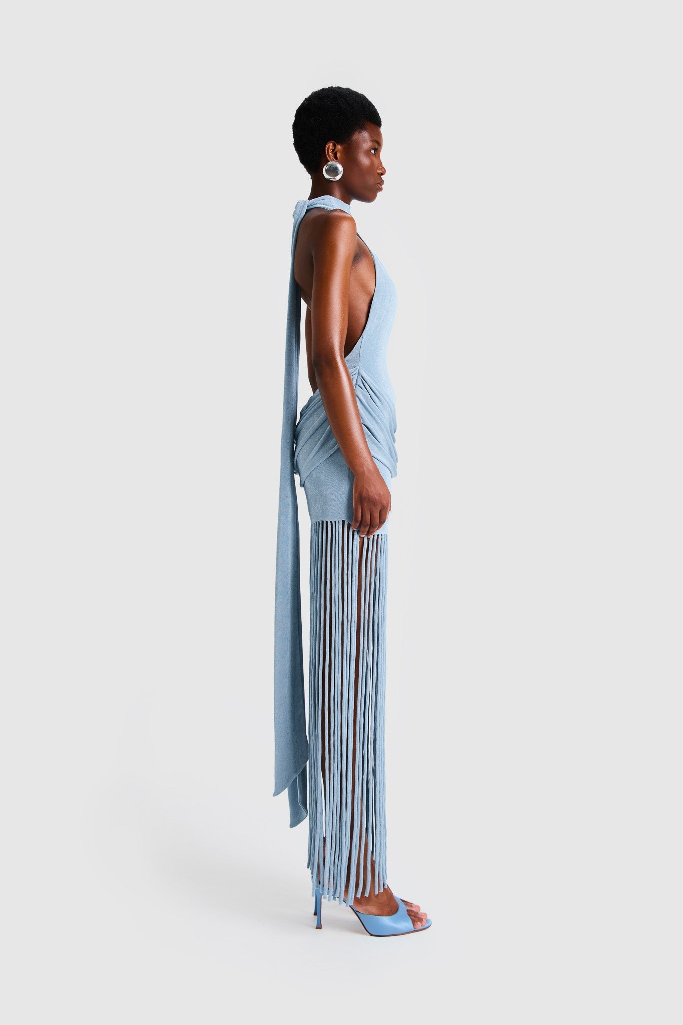 Full-length studio shot of a Black model wearing a sleeveless, high-neck halter dress in a deep red. The dress is form-fitting with ruching at the hips and features a dramatic, floor-length hem made of long, thick corded fringe.
