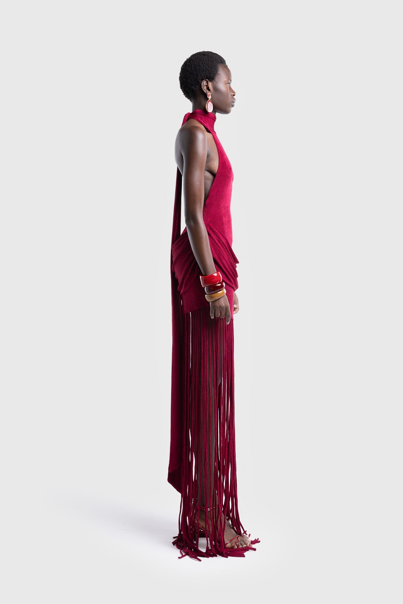 Full-length studio shot of a Black model wearing a sleeveless, high-neck halter dress in a deep red. The dress is form-fitting with ruching at the hips and features a dramatic, floor-length hem made of long, thick corded fringe.