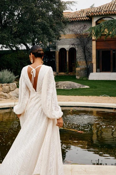 A woman poses in a garden wearing a long-sleeved, off-white maxi dress with a deep V-neck, voluminous bishop sleeves, a ruched waist, and a fully pleated skirt with a subtle wave pattern.