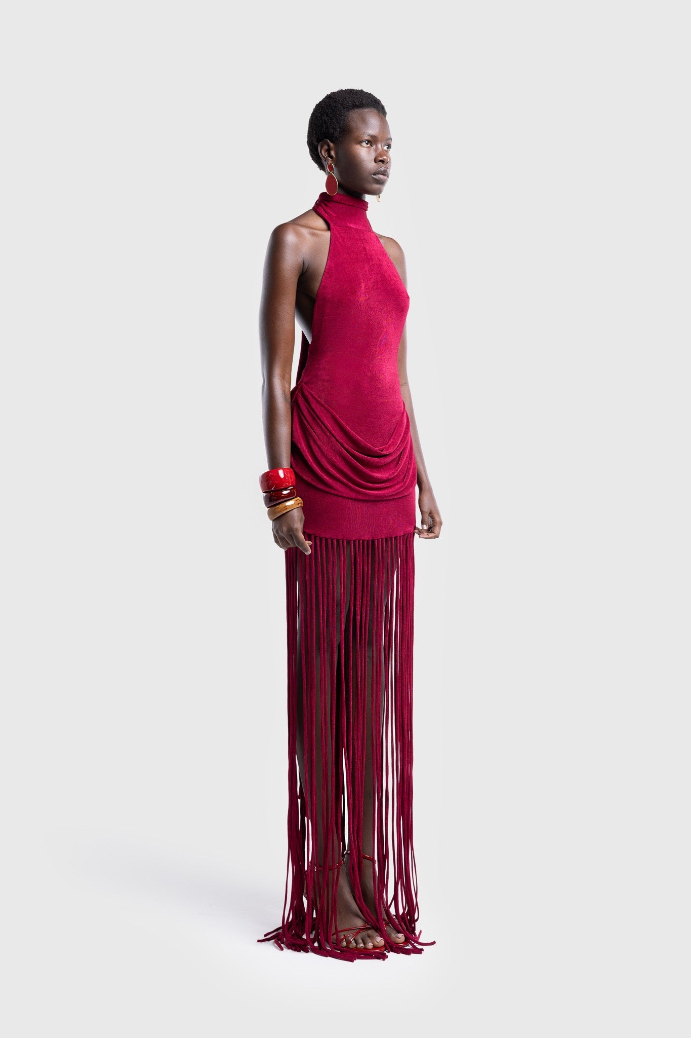 Full-length studio shot of a Black model wearing a sleeveless, high-neck halter dress in a deep red. The dress is form-fitting with ruching at the hips and features a dramatic, floor-length hem made of long, thick corded fringe.