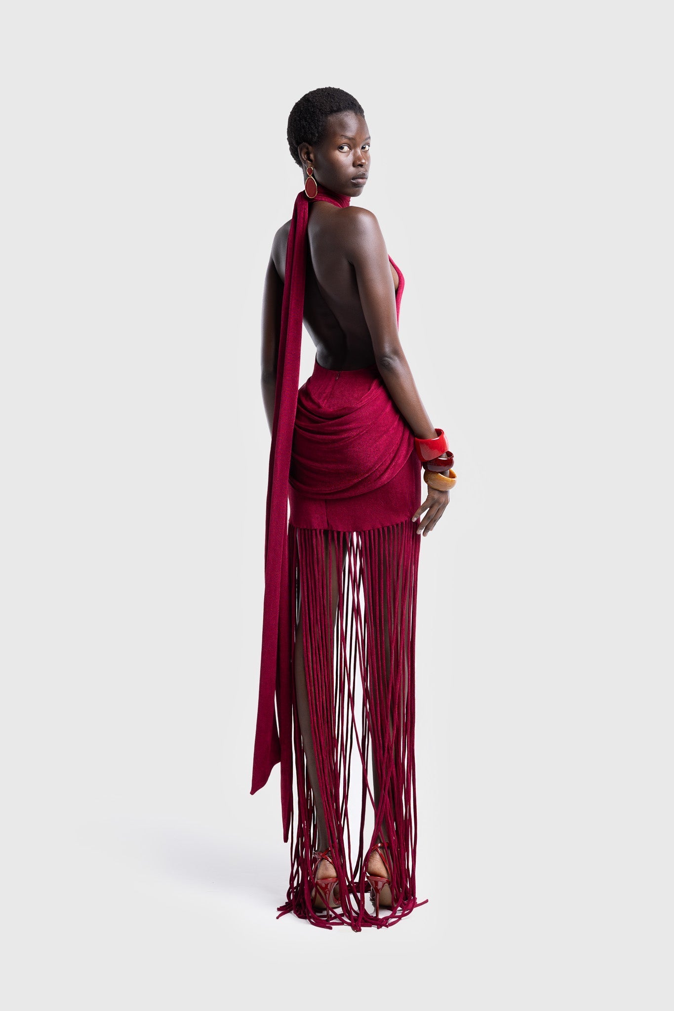 Full-length studio shot of a Black model wearing a sleeveless, high-neck halter dress in a deep red. The dress is form-fitting with ruching at the hips and features a dramatic, floor-length hem made of long, thick corded fringe.