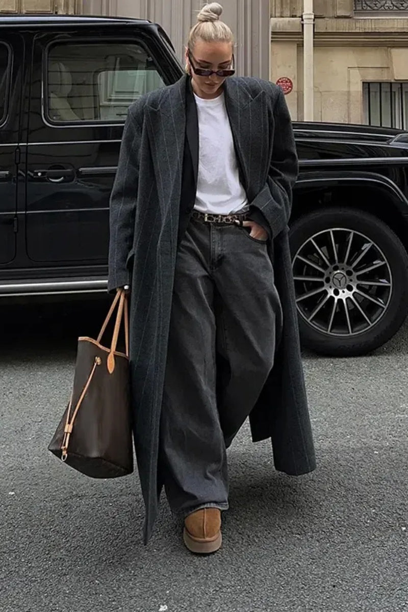 Grey - A woman walking on a city street wearing a long, oversized dark pinstripe maxi coat with structured shoulders and wide lapels.