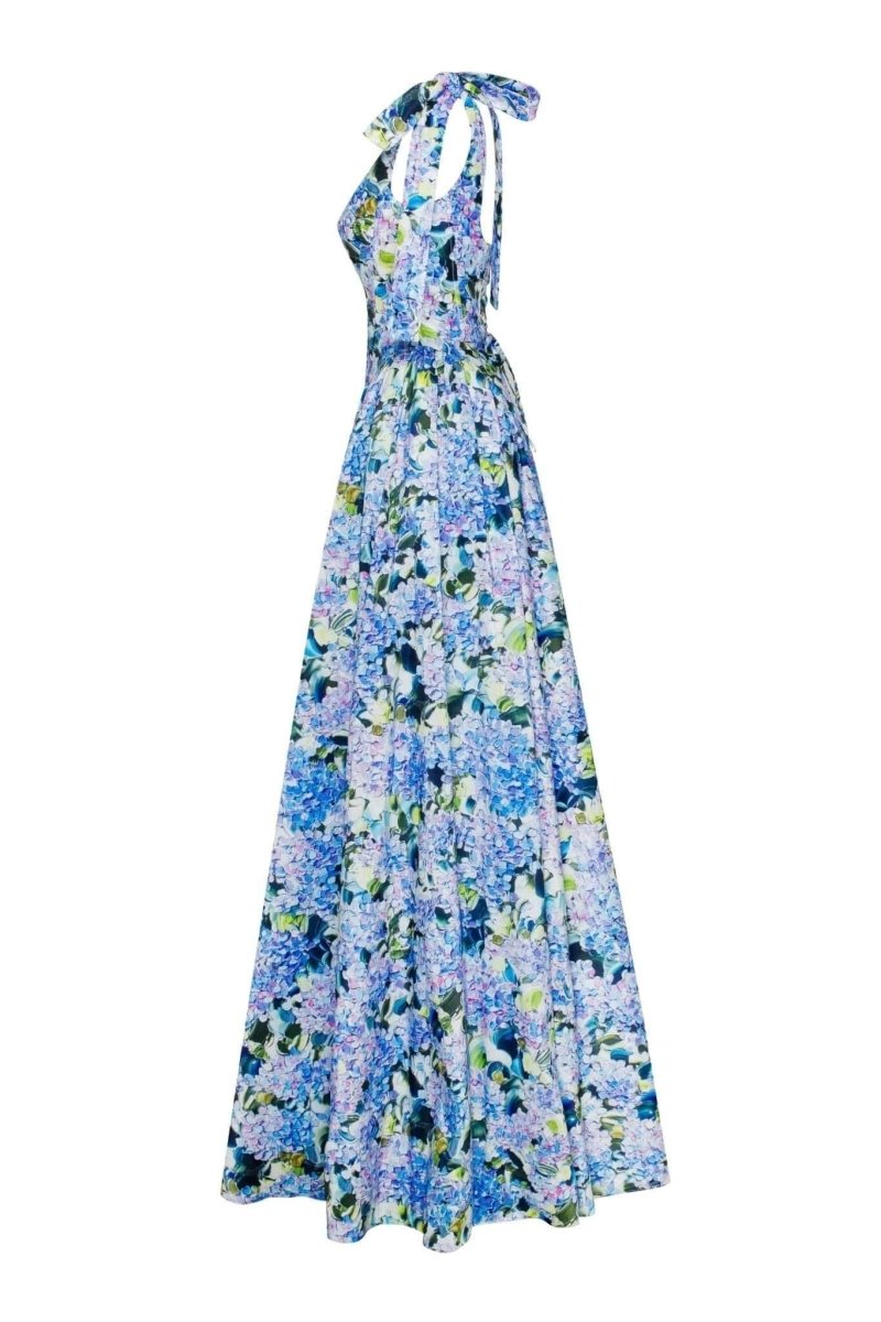 Full-length view of a model wearing an asymmetrical, tie-shoulder gown with a fitted bodice and a voluminous A-line skirt featuring a vibrant blue and purple hydrangea print, set against a white studio background.