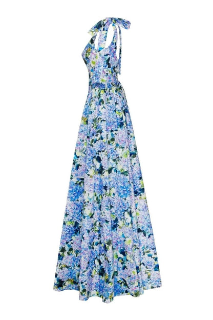 Full-length view of a model wearing an asymmetrical, tie-shoulder gown with a fitted bodice and a voluminous A-line skirt featuring a vibrant blue and purple hydrangea print, set against a white studio background.