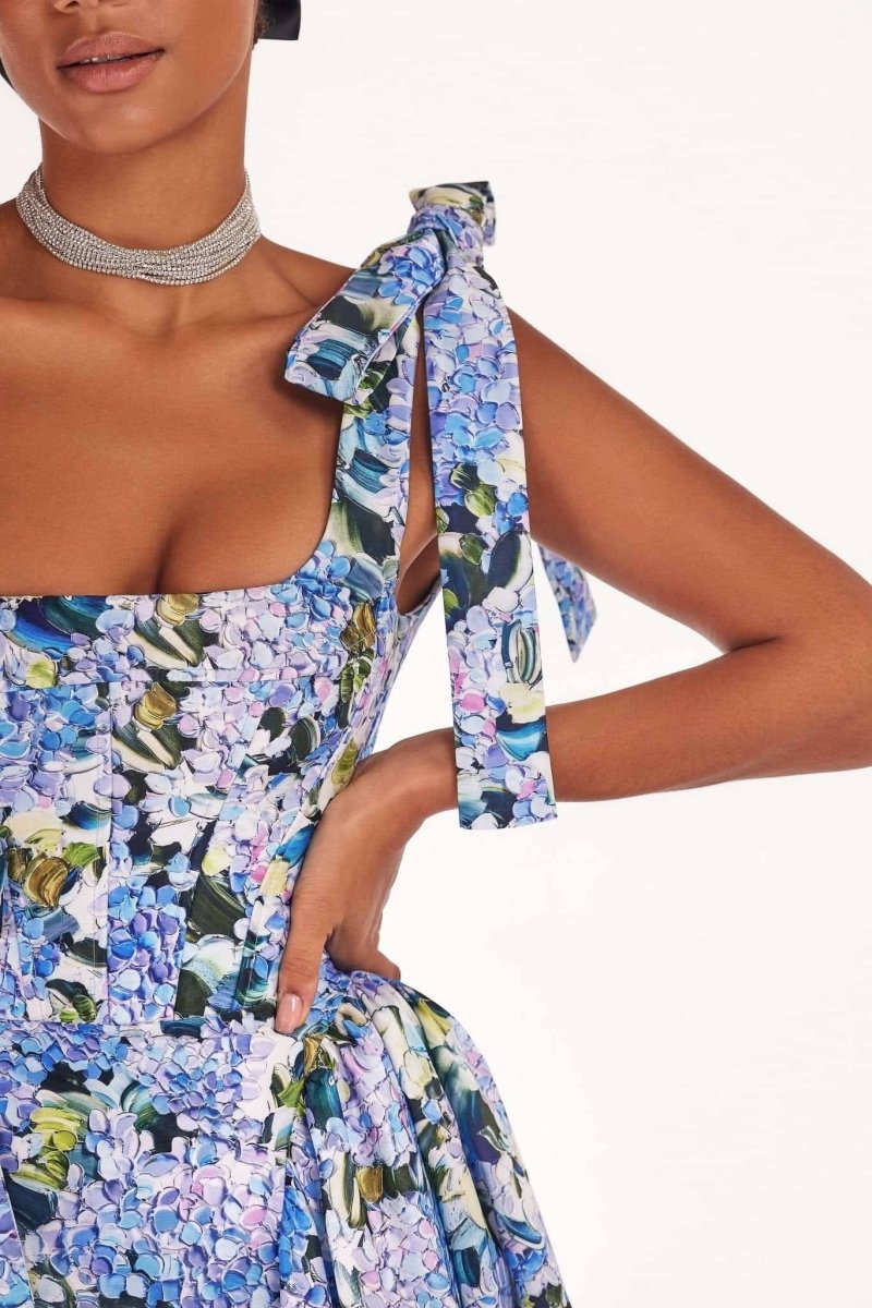 Full-length view of a model wearing an asymmetrical, tie-shoulder gown with a fitted bodice and a voluminous A-line skirt featuring a vibrant blue and purple hydrangea print, set against a white studio background.