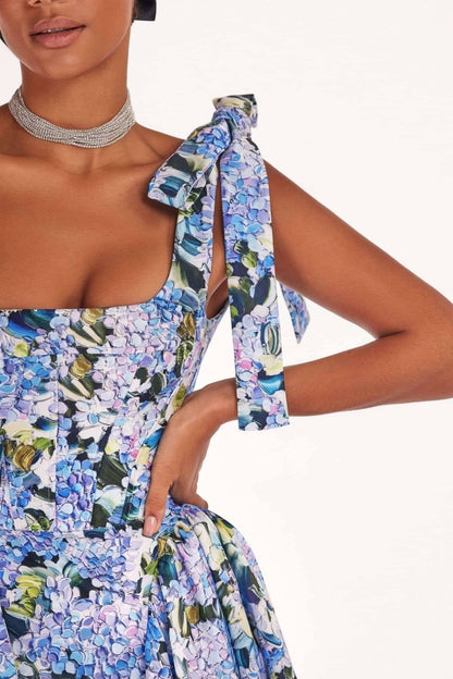 Full-length view of a model wearing an asymmetrical, tie-shoulder gown with a fitted bodice and a voluminous A-line skirt featuring a vibrant blue and purple hydrangea print, set against a white studio background.
