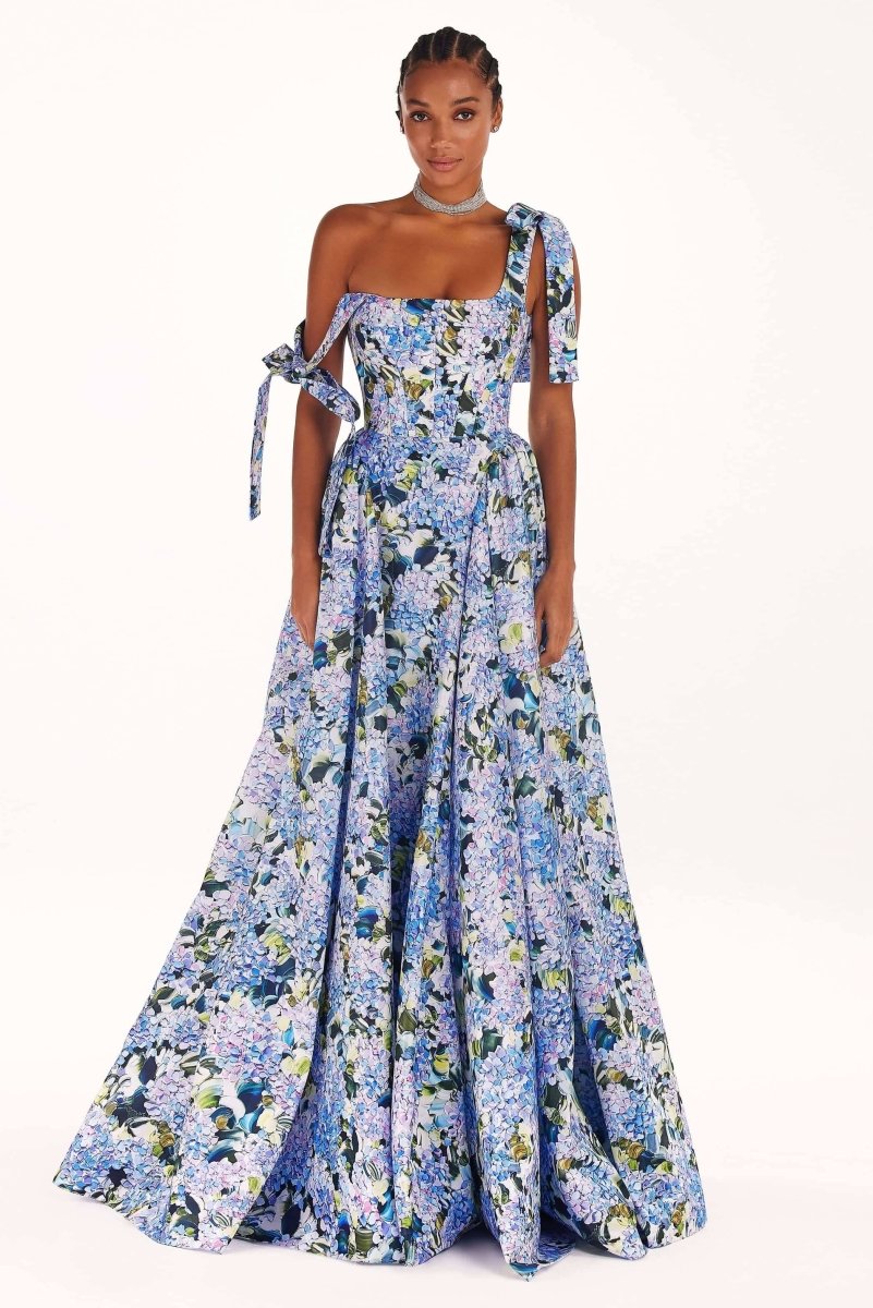 Full-length view of a model wearing an asymmetrical, tie-shoulder gown with a fitted bodice and a voluminous A-line skirt featuring a vibrant blue and purple hydrangea print, set against a white studio background.