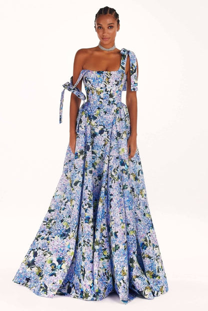 Full-length view of a model wearing an asymmetrical, tie-shoulder gown with a fitted bodice and a voluminous A-line skirt featuring a vibrant blue and purple hydrangea print, set against a white studio background.