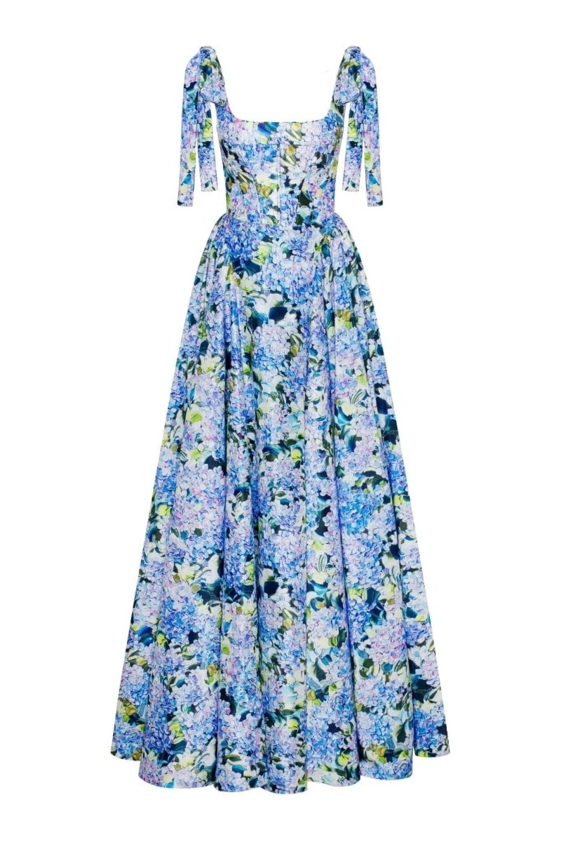 Full-length view of a model wearing an asymmetrical, tie-shoulder gown with a fitted bodice and a voluminous A-line skirt featuring a vibrant blue and purple hydrangea print, set against a white studio background.