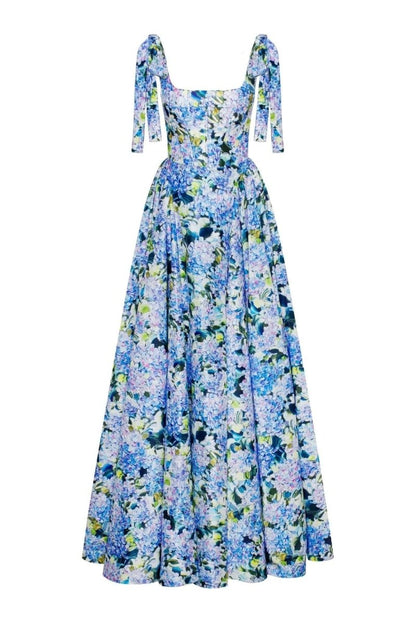 Full-length view of a model wearing an asymmetrical, tie-shoulder gown with a fitted bodice and a voluminous A-line skirt featuring a vibrant blue and purple hydrangea print, set against a white studio background.