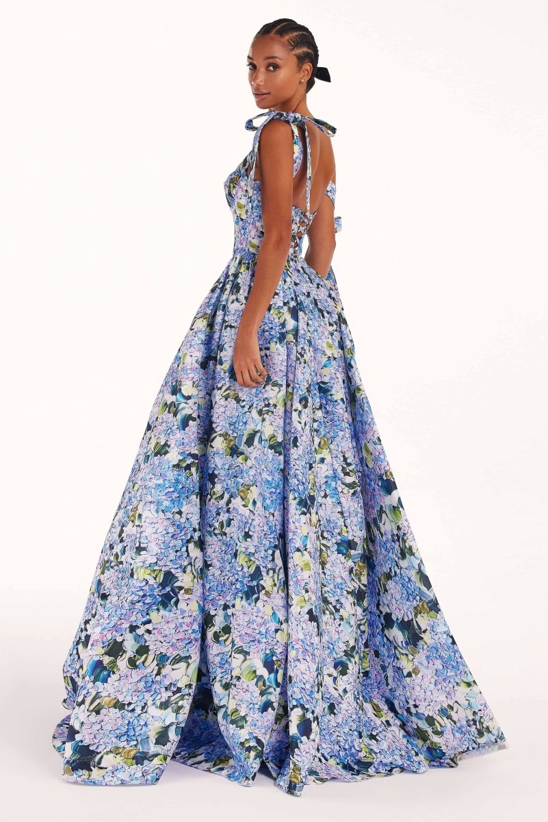 Full-length view of a model wearing an asymmetrical, tie-shoulder gown with a fitted bodice and a voluminous A-line skirt featuring a vibrant blue and purple hydrangea print, set against a white studio background.