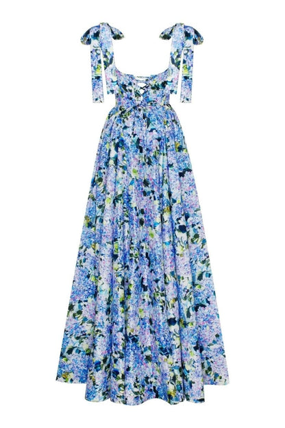 Full-length view of a model wearing an asymmetrical, tie-shoulder gown with a fitted bodice and a voluminous A-line skirt featuring a vibrant blue and purple hydrangea print, set against a white studio background.