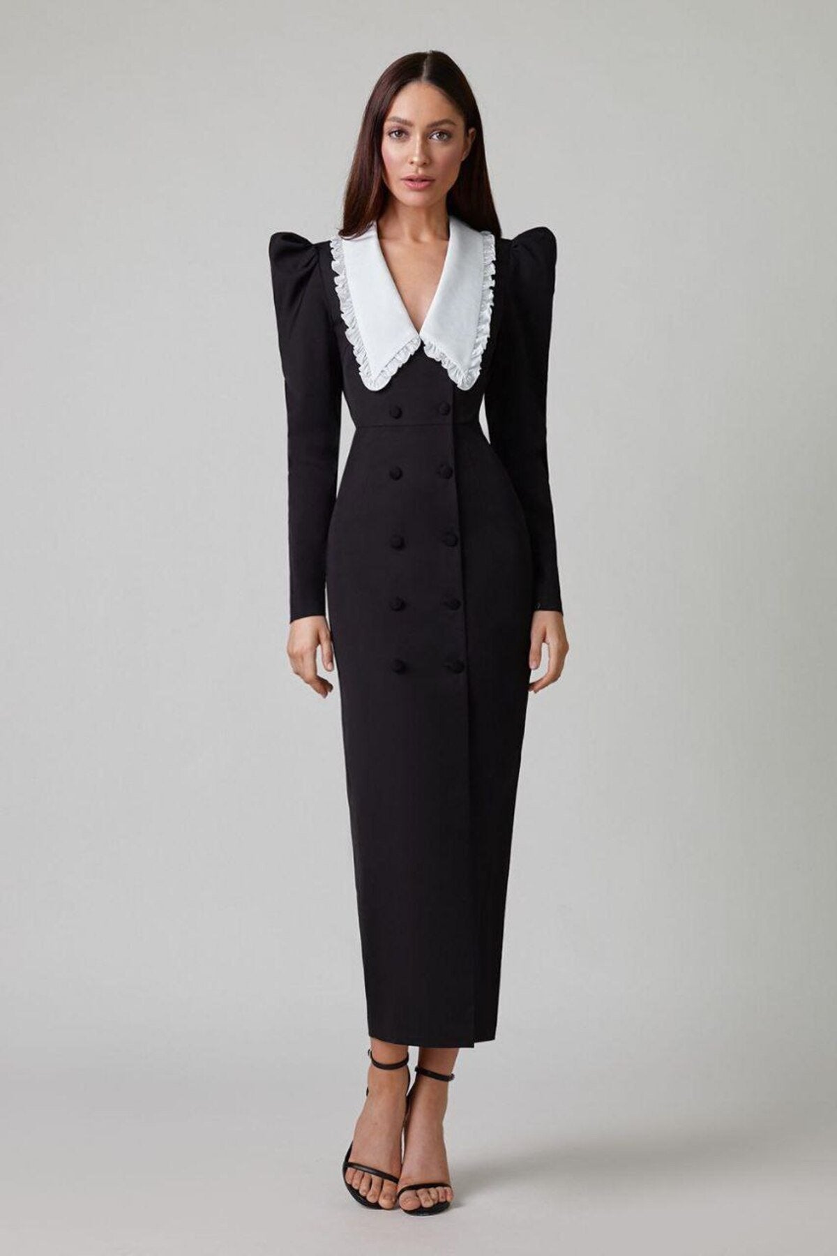 A woman models a long-sleeved, form-fitting black midi dress featuring a double-breasted front, structured puff shoulders, and a large, contrasting white collar with a ruffle edge.