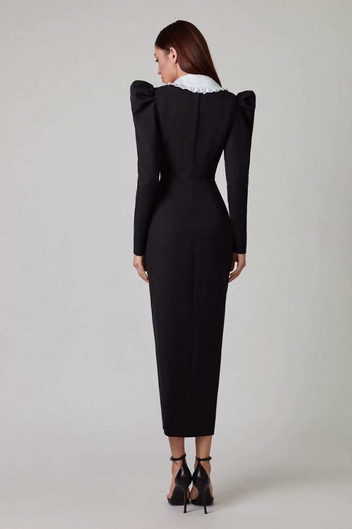 A woman models a long-sleeved, form-fitting black midi dress featuring a double-breasted front, structured puff shoulders, and a large, contrasting white collar with a ruffle edge.