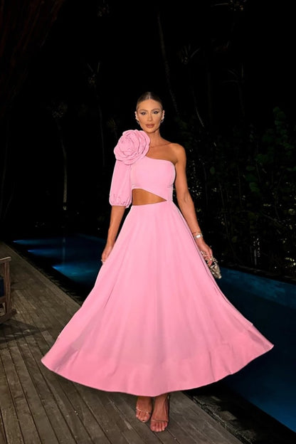 A woman viewed from the chest up, wearing a vibrant pink one-shoulder dress featuring a large, three-dimensional rose on the shoulder and a cutout on her side.
