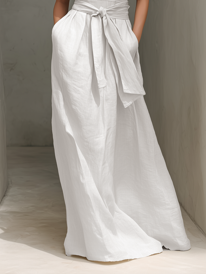 White - Woman wearing a minimalist sleeveless wide-leg jumpsuit with a deep V-neckline and a wide self-tie belt at the waist, posing in an architectural setting.