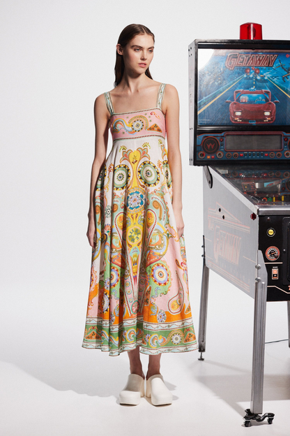 Multicolor - A model wearing a linen midi dress with a colorful kaleidoscope print, featuring a square neckline, wide straps, and an A-line skirt, standing next to a vintage pinball machine.
