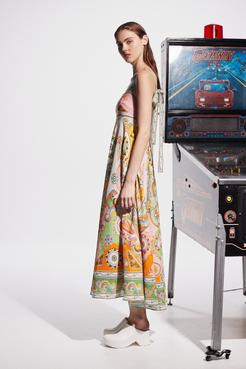 Multicolor - A model wearing a linen midi dress with a colorful kaleidoscope print, featuring a square neckline, wide straps, and an A-line skirt, standing next to a vintage pinball machine.
