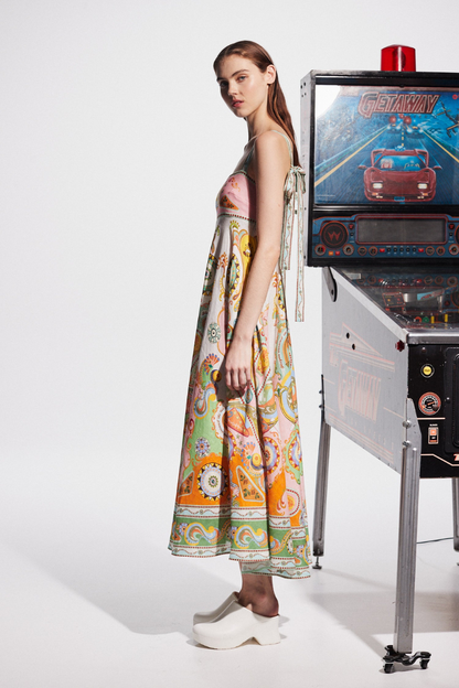 Multicolor - A model wearing a linen midi dress with a colorful kaleidoscope print, featuring a square neckline, wide straps, and an A-line skirt, standing next to a vintage pinball machine.