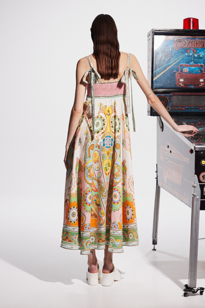 Multicolor - A model wearing a linen midi dress with a colorful kaleidoscope print, featuring a square neckline, wide straps, and an A-line skirt, standing next to a vintage pinball machine.