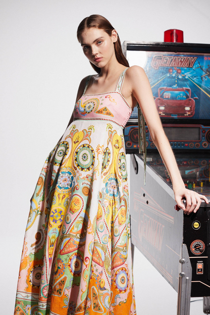 Multicolor - A model wearing a linen midi dress with a colorful kaleidoscope print, featuring a square neckline, wide straps, and an A-line skirt, standing next to a vintage pinball machine.