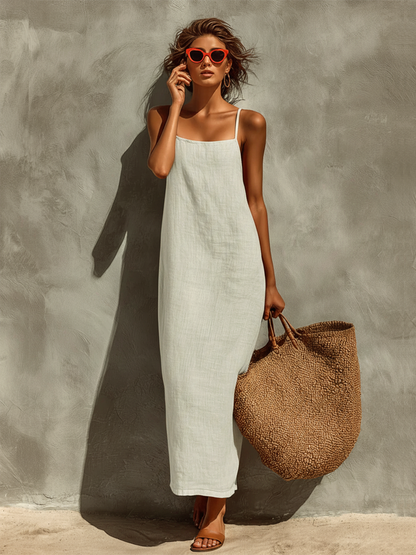 White - Woman wearing a textured maxi slip sundress with spaghetti straps, holding a large woven tote bag against a textured grey wall.