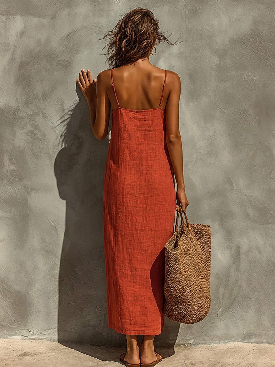White - Woman wearing a textured maxi slip sundress with spaghetti straps, holding a large woven tote bag against a textured grey wall.