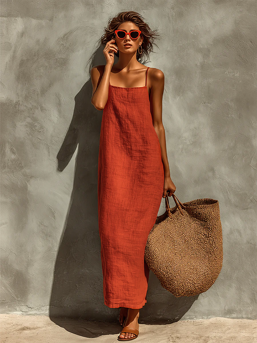 Red - Woman wearing a textured maxi slip sundress with spaghetti straps, holding a large woven tote bag against a textured grey wall.