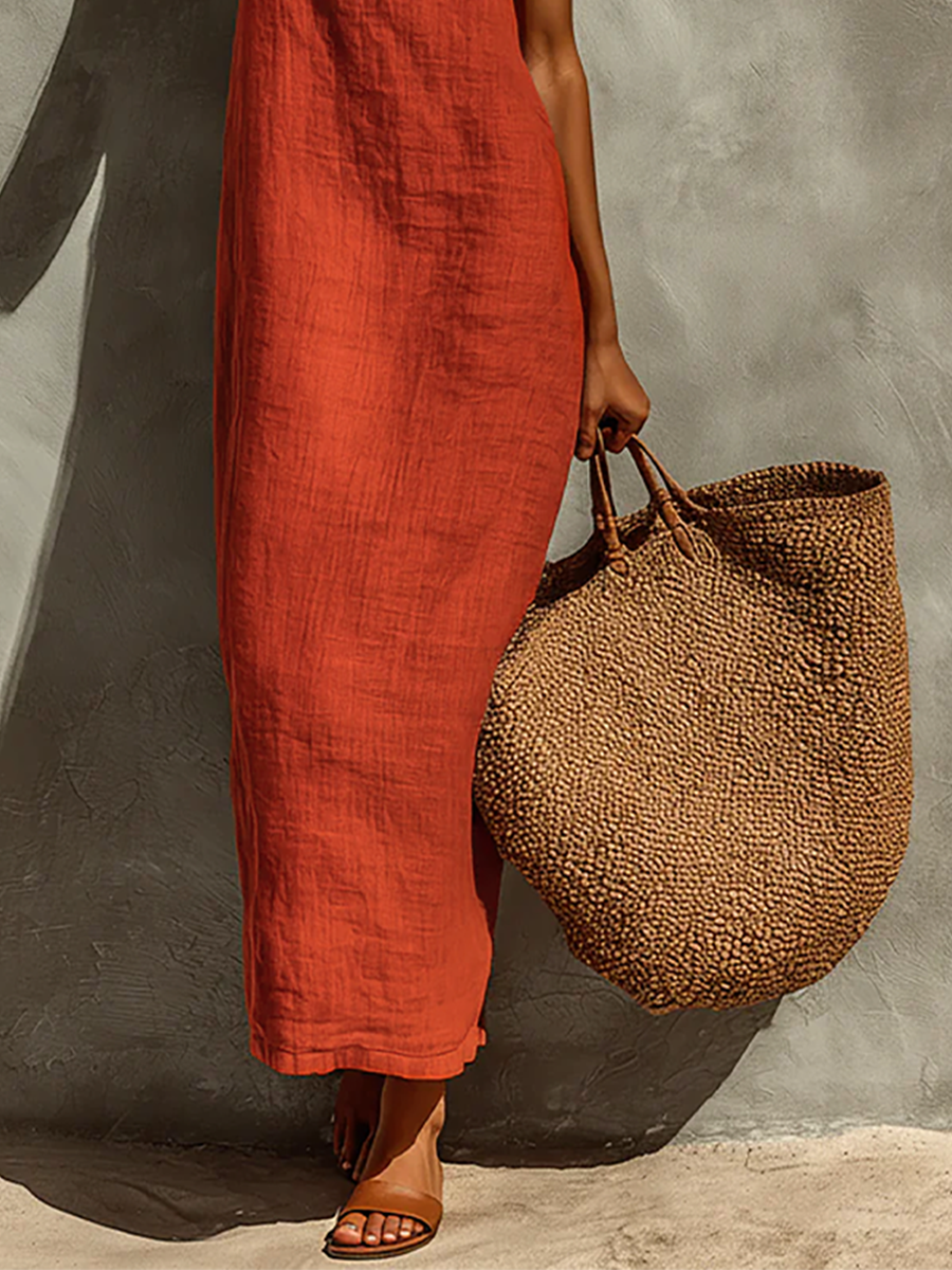 Red - Woman wearing a textured maxi slip sundress with spaghetti straps, holding a large woven tote bag against a textured grey wall.