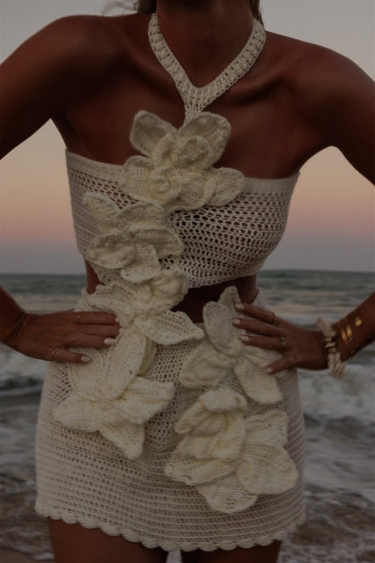 A woman on a beach wearing a crochet knit cutout mini dress with a halter neck and large 3D floral appliqués.