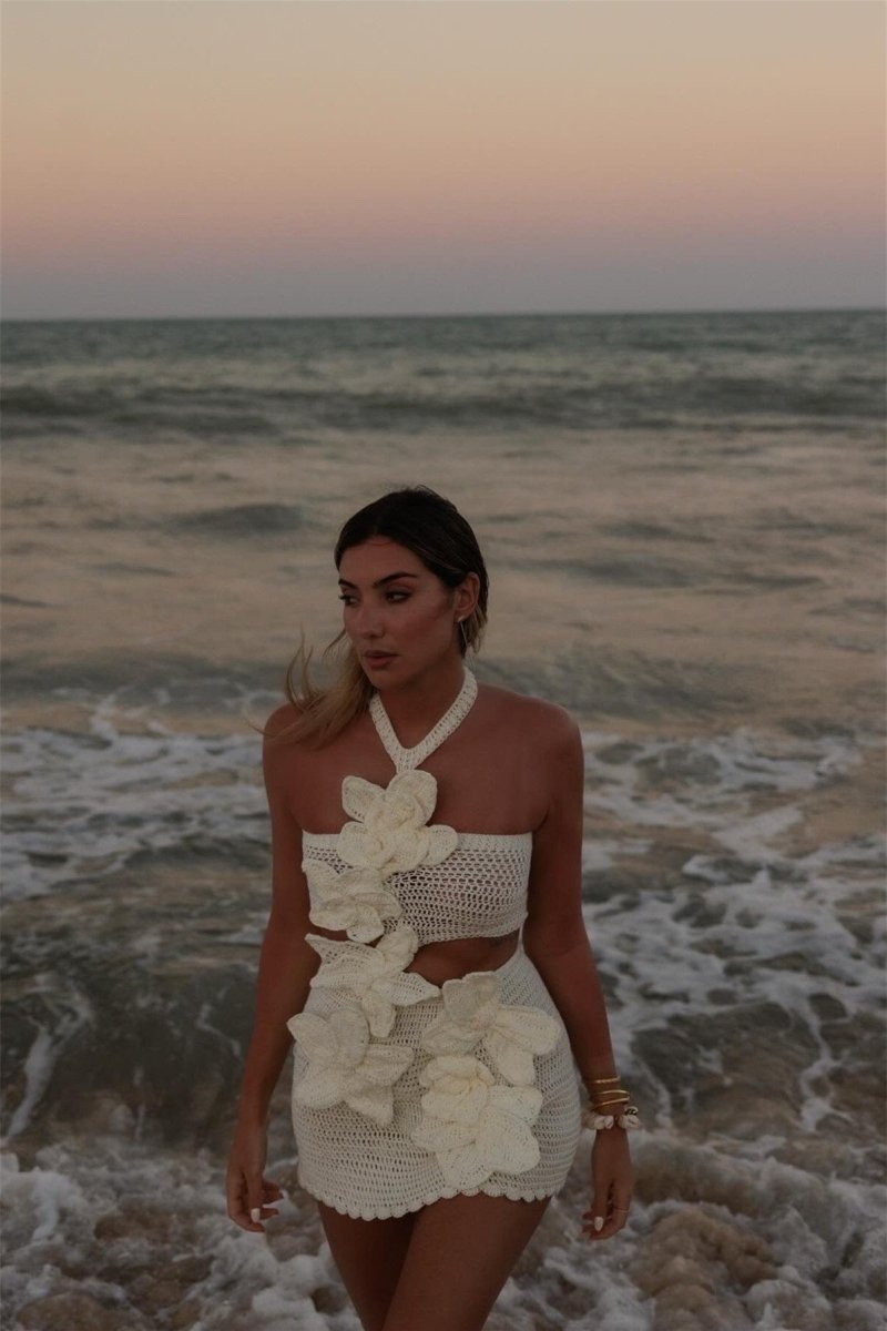 A woman on a beach wearing a form-fitting, cream crochet halter mini dress with a large side cut-out and oversized 3D floral appliqués.