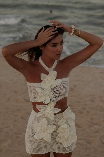 A woman on a beach wearing a form-fitting, cream crochet halter mini dress with a large side cut-out and oversized 3D floral appliqués.