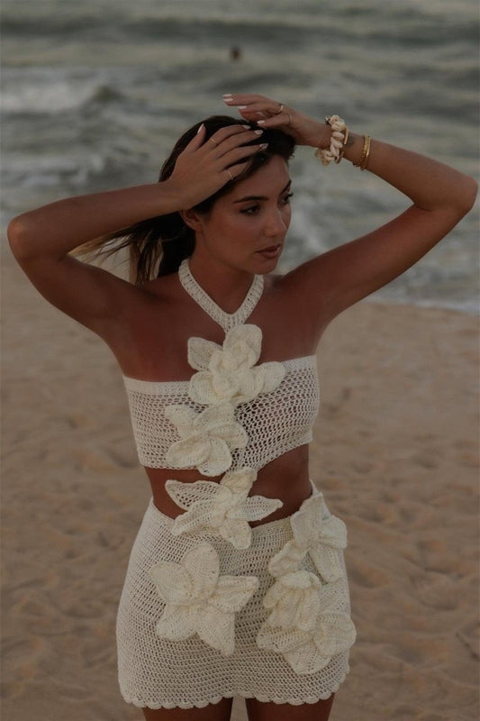 A woman on a sandy beach wears a cream-colored crochet halter top and matching mini skirt, connected by a cascade of 3D floral appliqués that create a dramatic waist cutout.