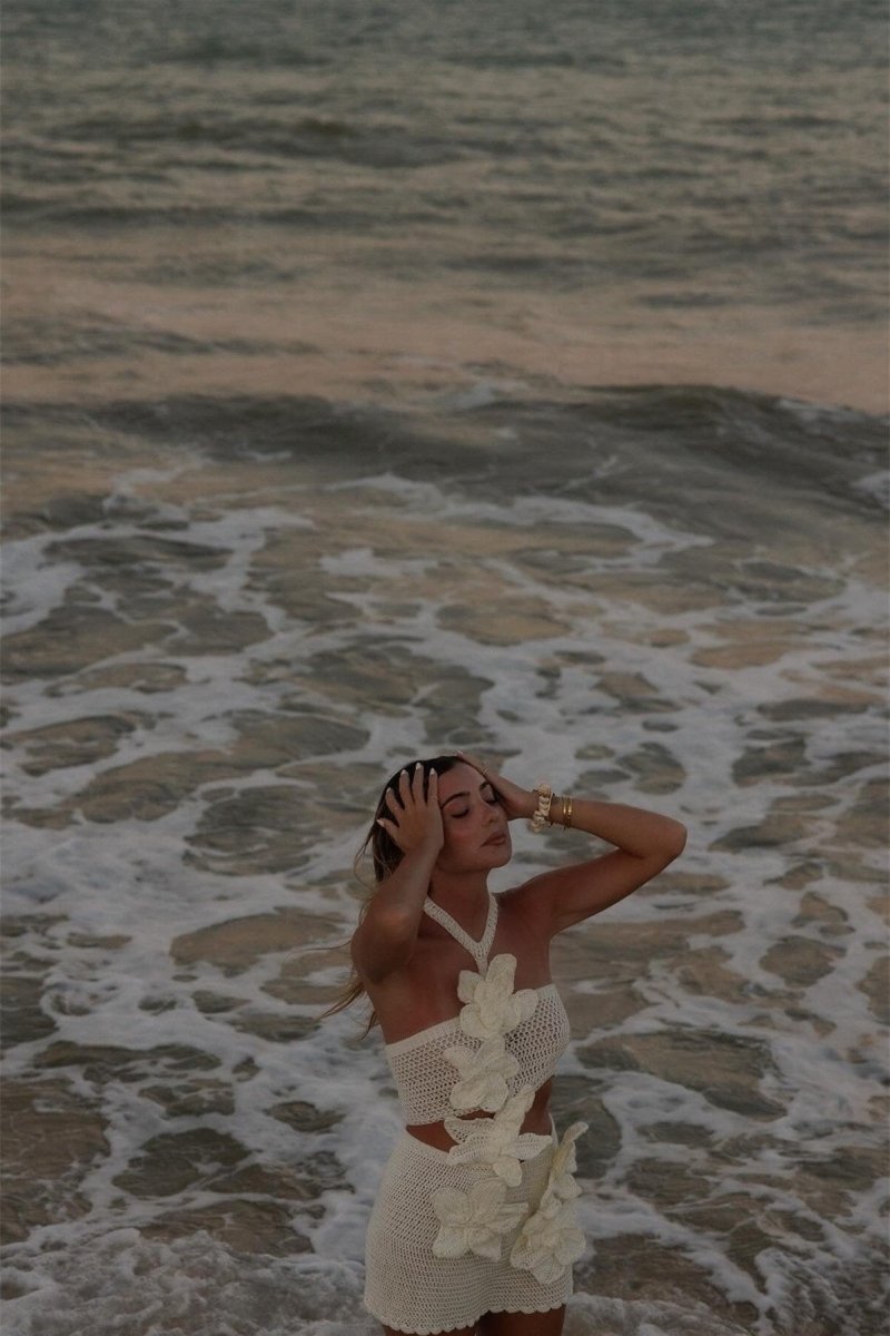 A woman on a beach wearing a form-fitting, cream crochet halter mini dress with a large side cut-out and oversized 3D floral appliqués.