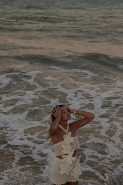 A woman on a beach wearing a form-fitting, cream crochet halter mini dress with a large side cut-out and oversized 3D floral appliqués.