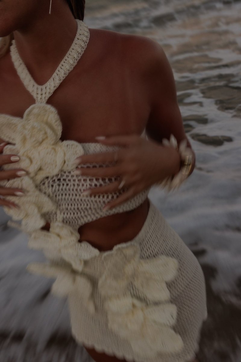 A woman on a beach wearing a crochet knit cutout mini dress with a halter neck and large 3D floral appliqués.