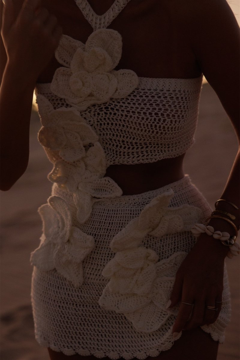 A woman on a beach wearing a form-fitting, cream crochet halter mini dress with a large side cut-out and oversized 3D floral appliqués.