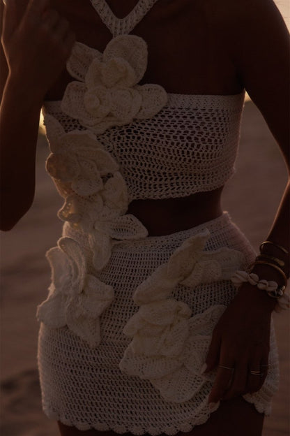 A woman on a beach wearing a crochet knit cutout mini dress with a halter neck and large 3D floral appliqués.