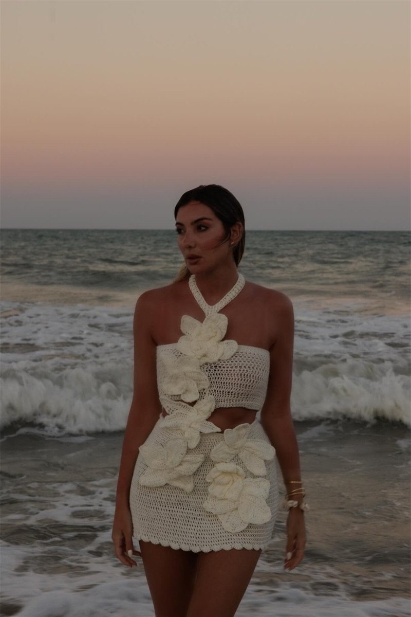 A woman on a beach wearing a crochet knit cutout mini dress with a halter neck and large 3D floral appliqués.